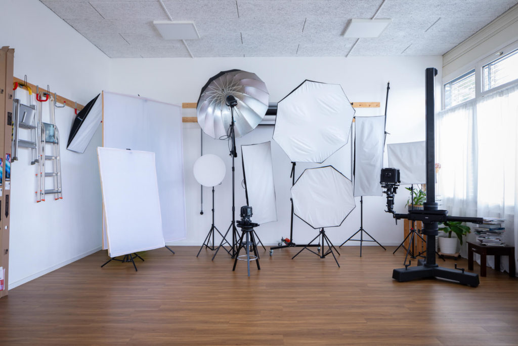 location-studio-photo-video-lausanne-equipé-015 - Location de Studio équipé pour la Photo et la Vidéo Un studio photo avec un sol en bois, plusieurs grandes lampes de type softbox, des parapluies, des appareils photo sur trépieds, des réflecteurs et des fonds posés contre un mur blanc près d'une fenêtre avec de fins rideaux.
